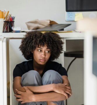 A woman appears overwhelmed, sitting under an office desk in a work environment.