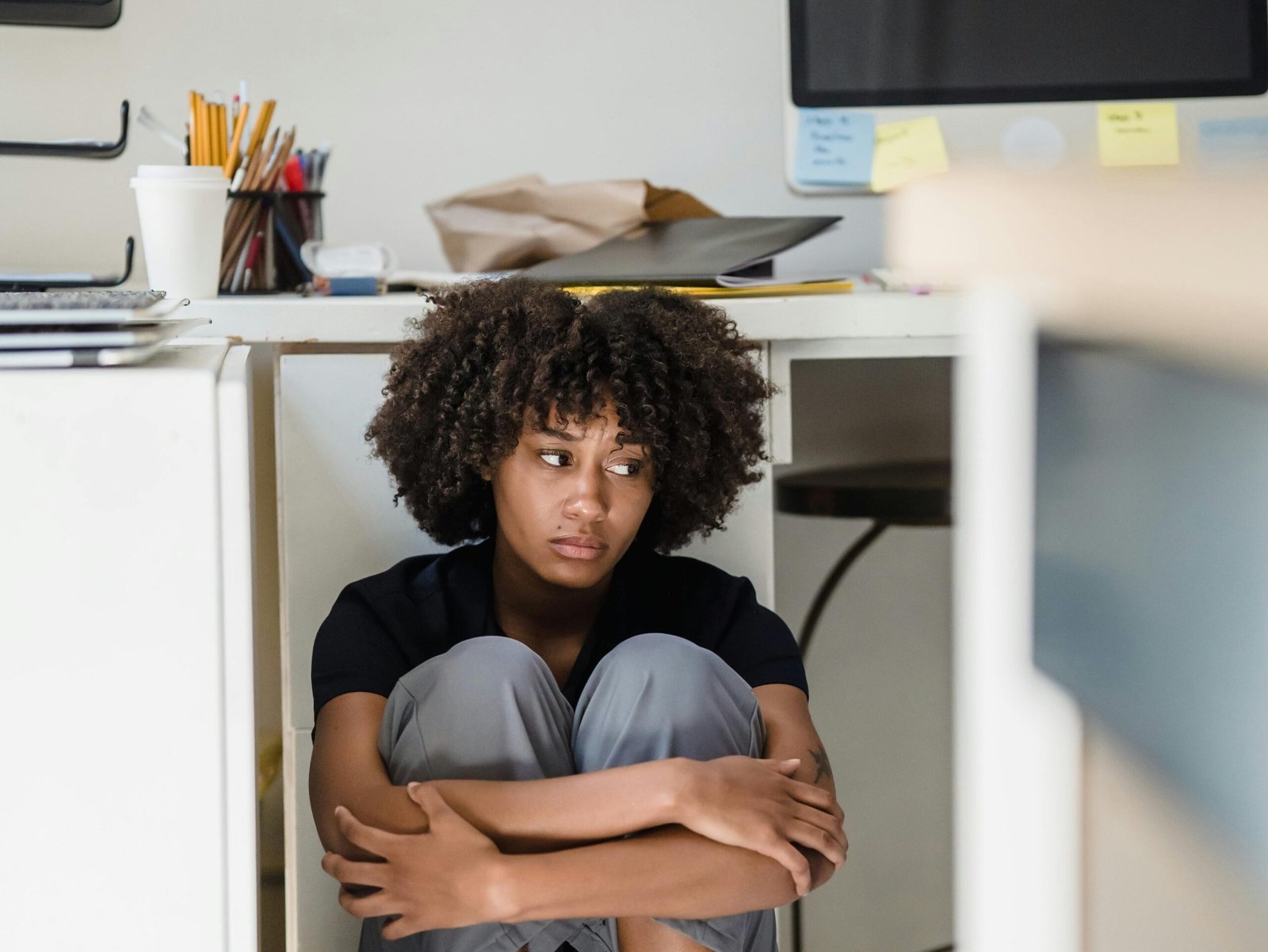 A woman appears overwhelmed, sitting under an office desk in a work environment.