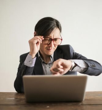 Young frowning man in suit and glasses looking at wristwatch while waiting for appointment sitting at desk with laptop