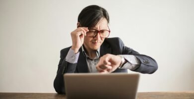 Young frowning man in suit and glasses looking at wristwatch while waiting for appointment sitting at desk with laptop