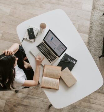 From above of young woman with long dark hair in casual clothes working at table and browsing netbook while sitting in modern workplace and touching hair