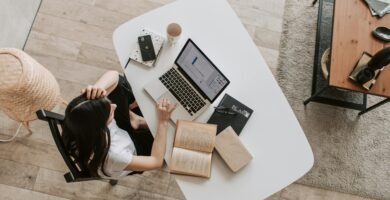 From above of young woman with long dark hair in casual clothes working at table and browsing netbook while sitting in modern workplace and touching hair