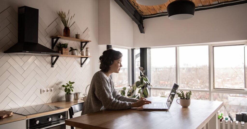 Side view of young female in casual clothes typing message on laptop while sitting at wooden table in kitchen