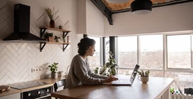 Side view of young female in casual clothes typing message on laptop while sitting at wooden table in kitchen