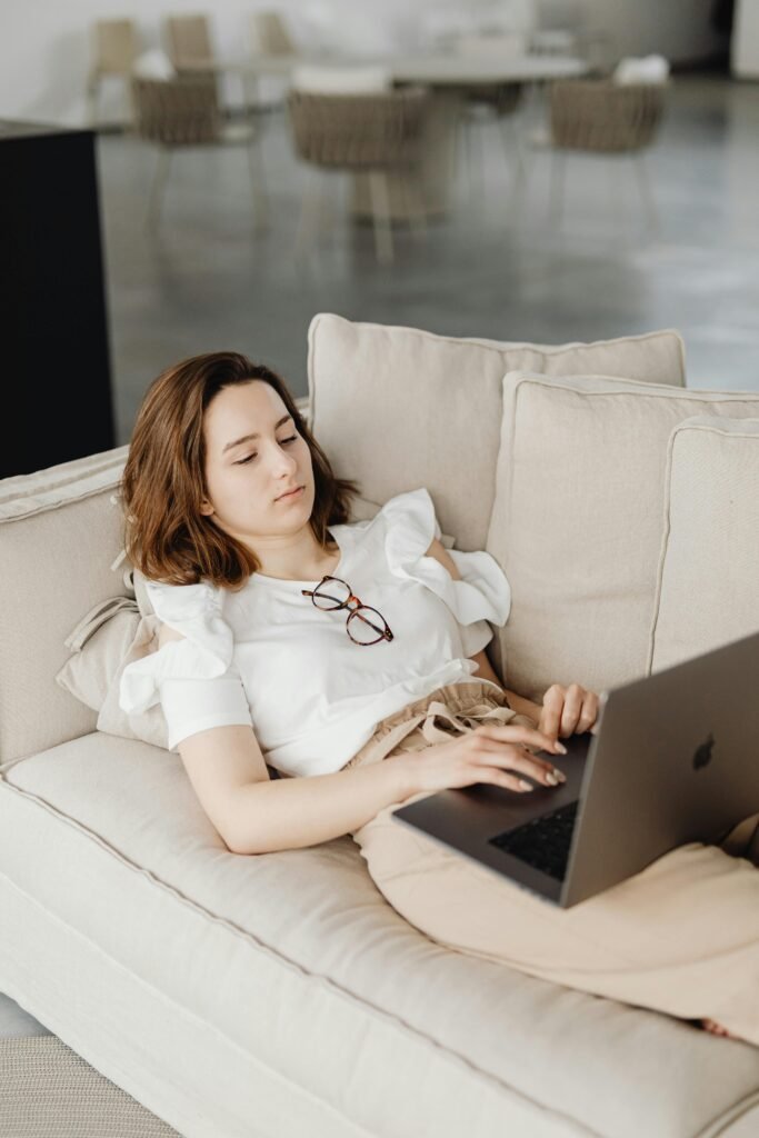 Woman lying on a couch working remotely on a laptop, embodying a comfortable home office setting.
